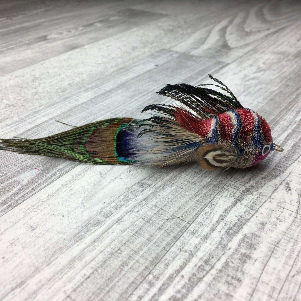 This is a Pretty Fly Fish Teaser Wand Cat Toy Replacement Lure by Catboutique. The fish is made from deer hair, and it has a peacock eye feather for a tail. The fish has feathers for fins and dorsal fin. There is a ringlet where the fish's mouth should be. The fish is blue, red, white and black. This lure is meant to engage at the cat's hunting instincts like prowling, pawing, and pouncing. 