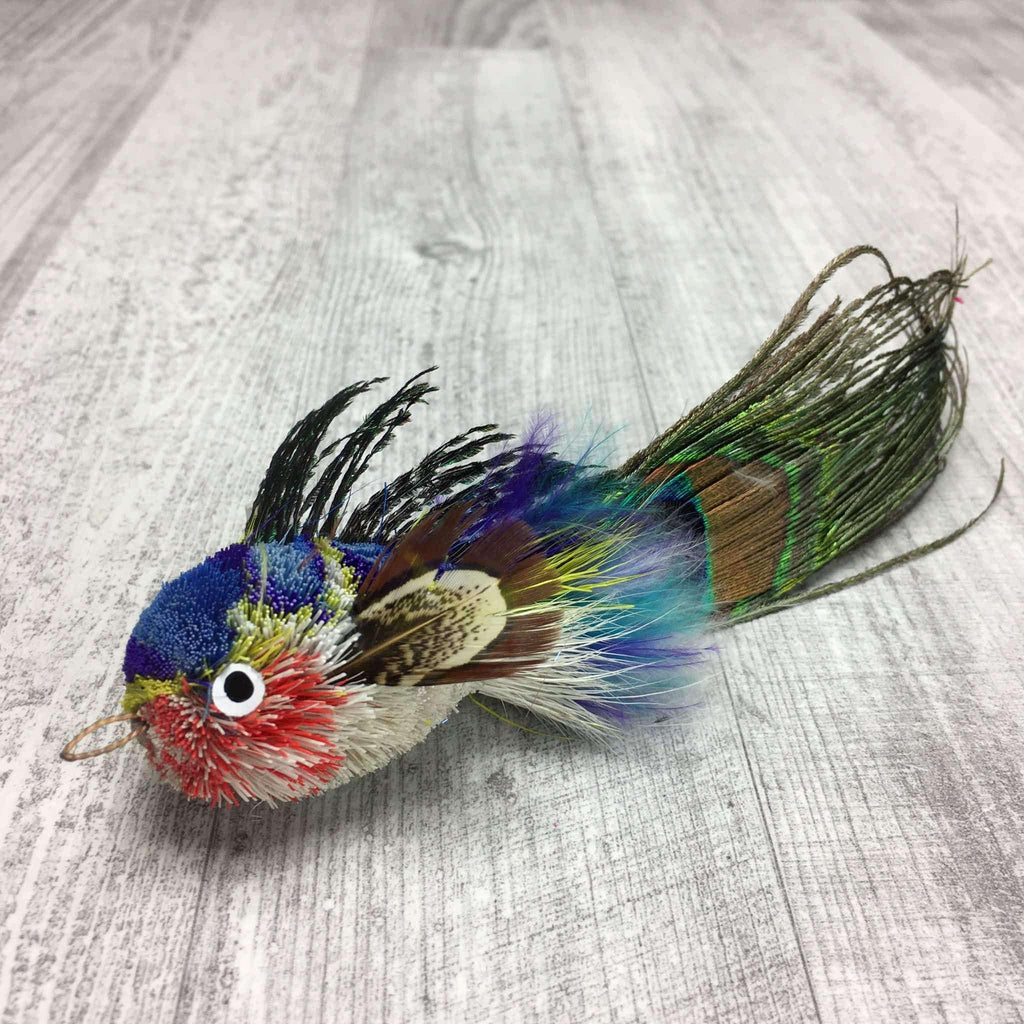 This is a Pretty Fly Fish Teaser Wand Cat Toy Replacement Lure by Catboutique. The fish is made from deer hair, and it has a peacock eye feather for a tail. The fish has feathers for fins and dorsal fin. There is a ringlet where the fish's mouth should be. The fish is red, blue, yellow, and white. This lure is meant to engage at the cat's hunting instincts like prowling, pawing, and pouncing.  