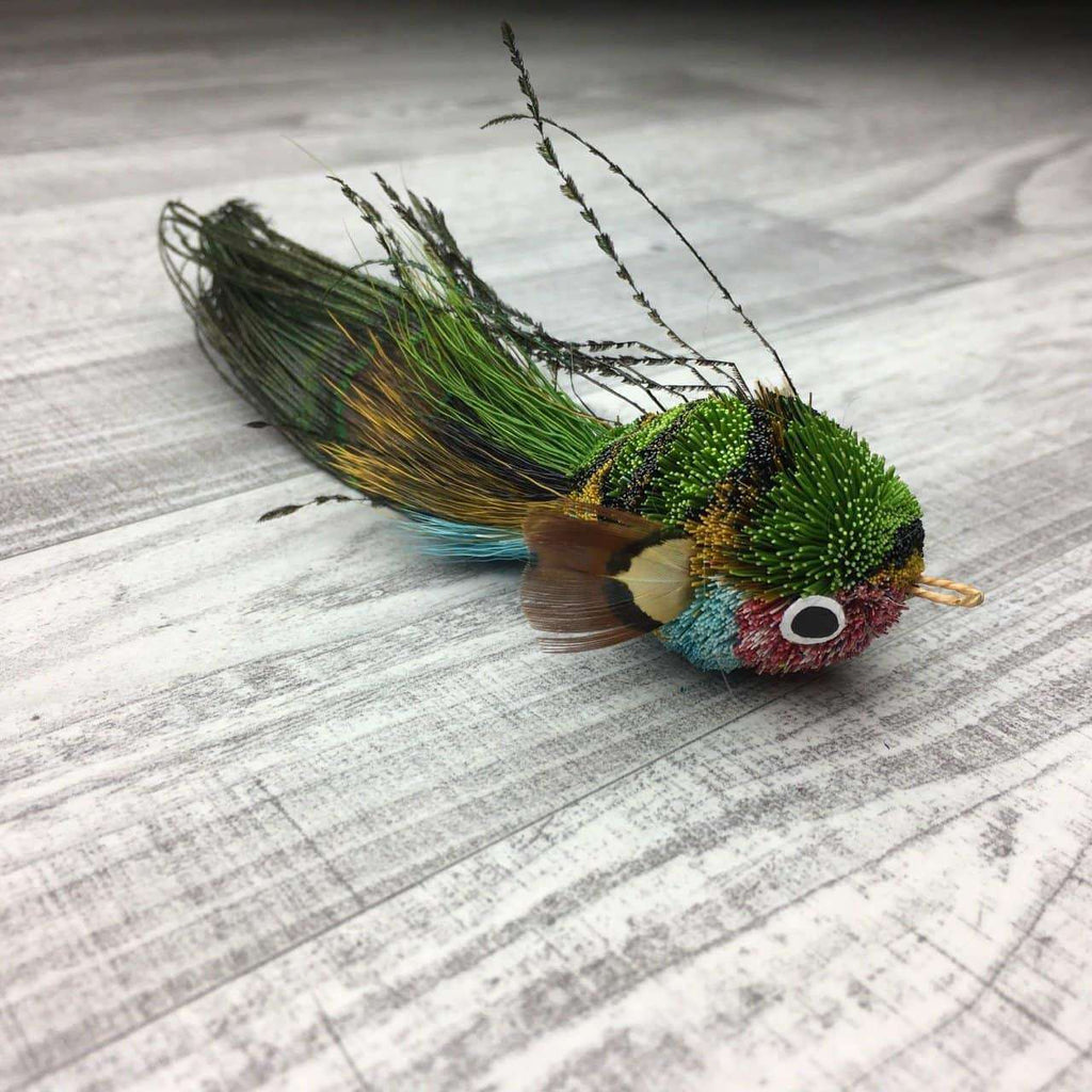This is a Pretty Fly Fish Teaser Wand Cat Toy Replacement Lure by Catboutique. The fish is made from deer hair, and it has a peacock eye feather for a tail. The fish has feathers for fins and dorsal fin. There is a ringlet where the fish's mouth should be. The fish is blue, red, green, and black. This lure is meant to engage at the cat's hunting instincts like prowling, pawing, and pouncing.  