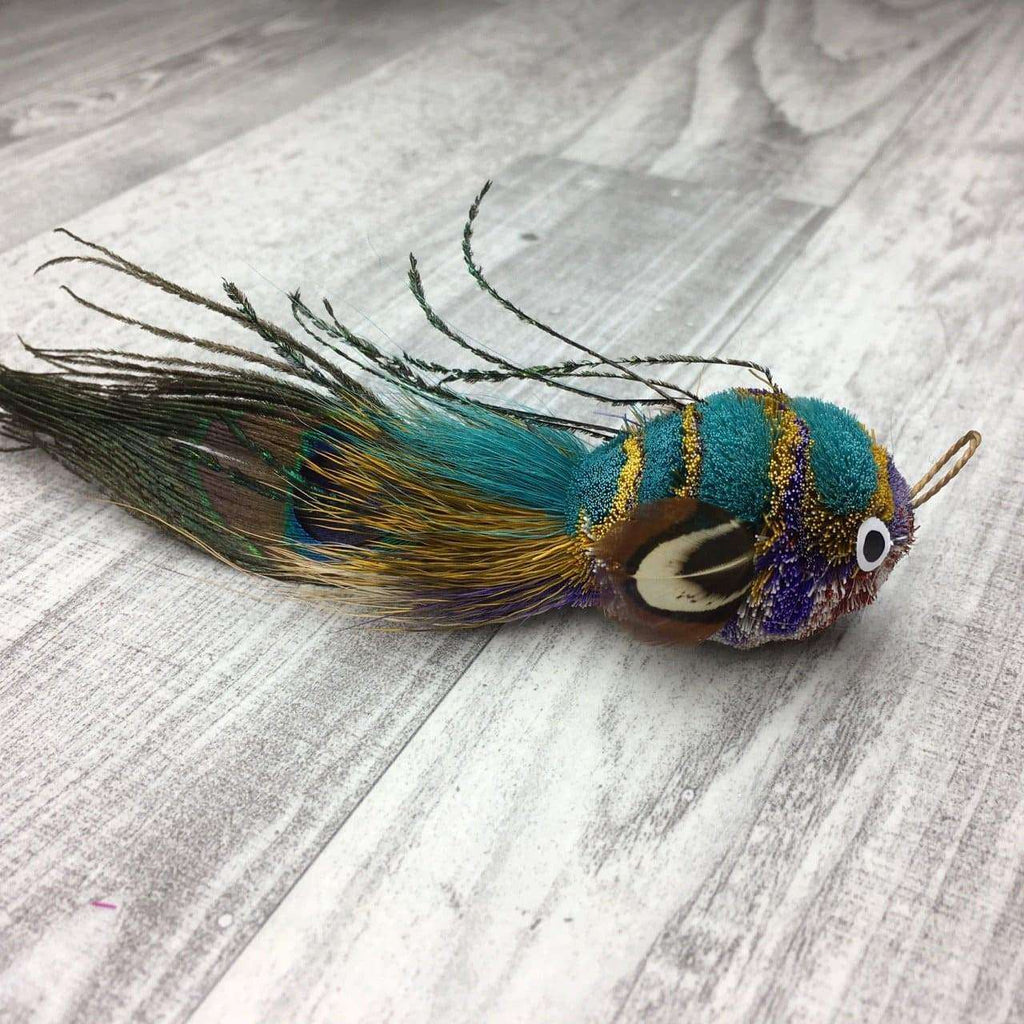 This is a Pretty Fly Fish Teaser Wand Cat Toy Replacement Lure by Catboutique. The fish is made from deer hair, and it has a peacock eye feather for a tail. The fish has feathers for fins and dorsal fin. There is a ringlet where the fish's mouth should be. The fish is turquoise, gold, purple, and pink. This lure is meant to engage at the cat's hunting instincts like prowling, pawing, and pouncing.  